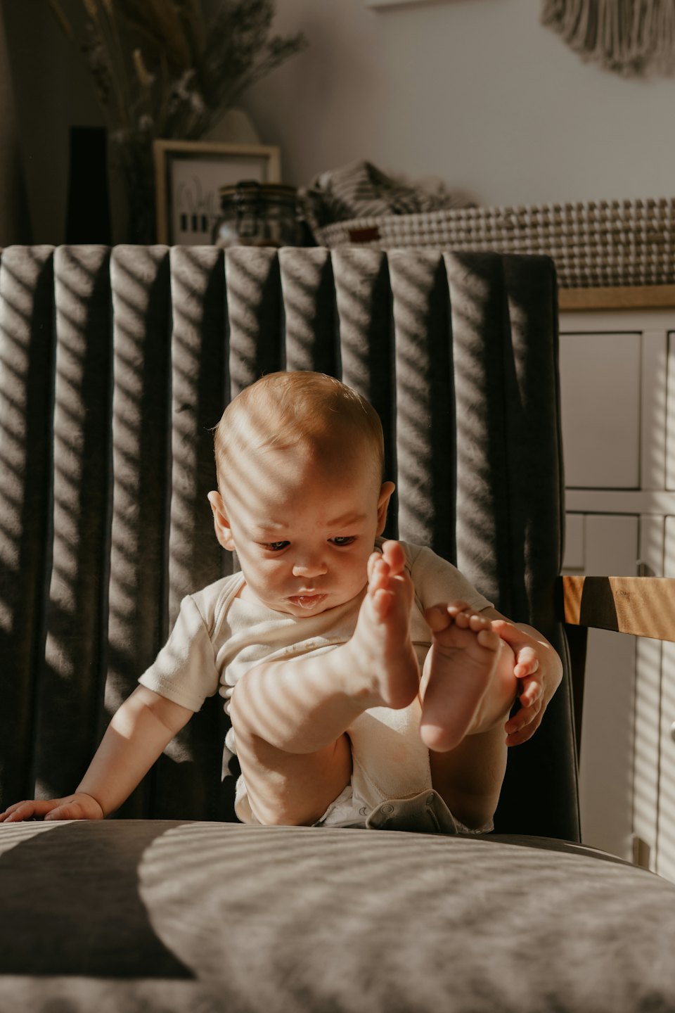 A baby wearing a white onesie sits on a gray chair playing with their feet.